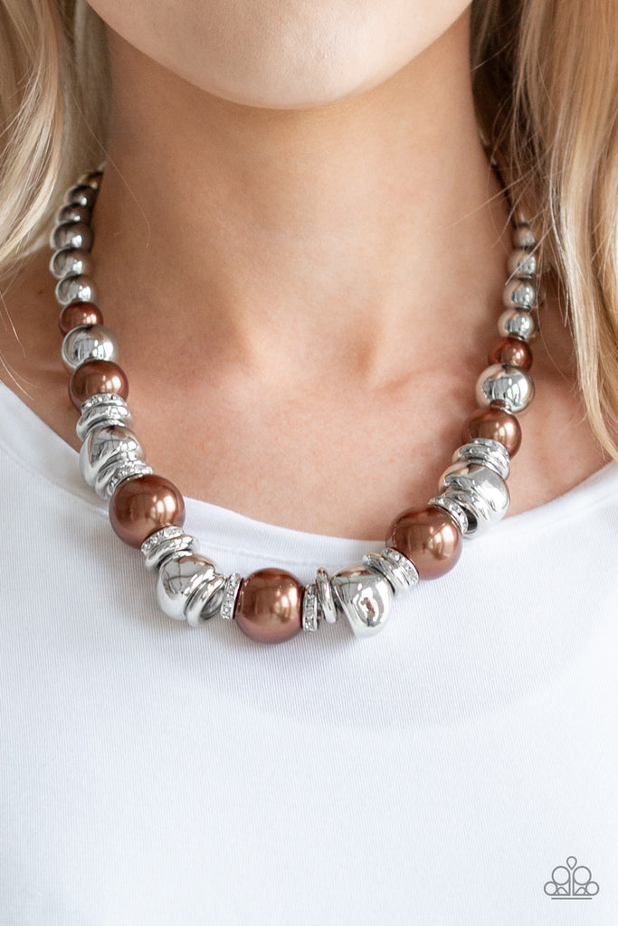 A collection of oversized brown pearls, silver rings, and asymmetrical silver beads are threaded along an invisible wire below the collar. White rhinestone encrusted rings are sprinkled between the refined beading for a statement-making finish. Features an adjustable clasp closure.  Sold as one individual necklace. Includes one pair of matching earrings.
