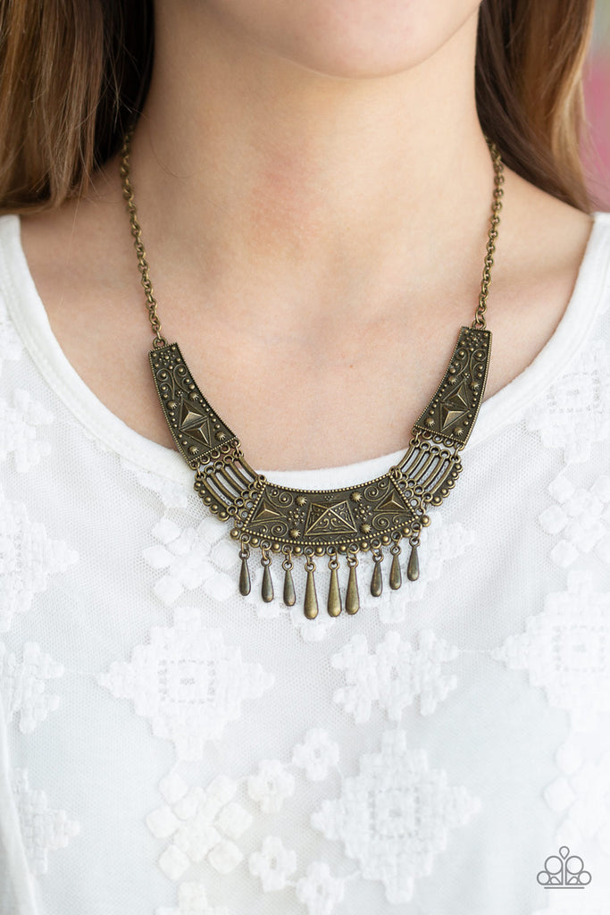 Embossed and studded in tribal inspired details, glistening brass plates connect below the collar in a fierce fashion. Dainty teardrop brass beads drip from the center of the pendant, creating a delicate fringe. Features an adjustable clasp closure.  Sold as one individual necklace. Includes one pair of matching earrings.