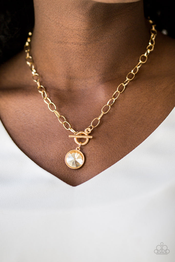 A dramatic golden gem swings from the bottom of a shiny gold chain, creating a blinding pendant below the collar. Features a toggle closure.  Sold as one individual necklace. Includes one pair of matching earrings.