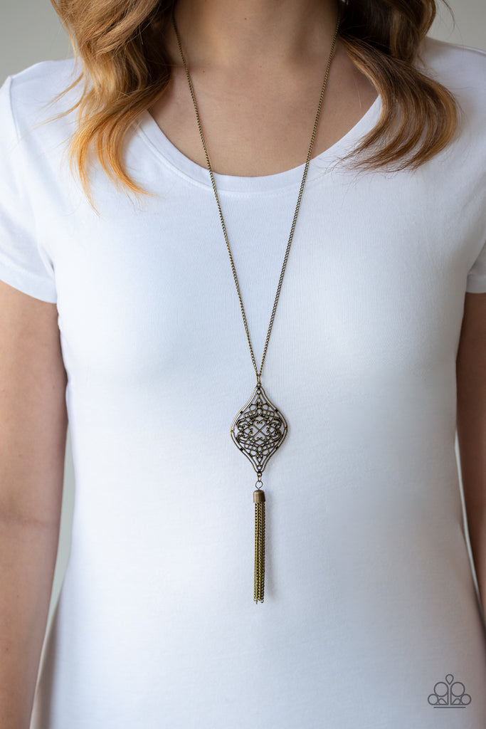 Swirling with whimsical filigree detail, an ornate brass frame swings from the bottom of a lengthened brass chain. A shimmery chain tassel is added for a wanderlust flair. Features an adjustable clasp closure.  Sold as one individual necklace. Includes one pair of matching earrings.