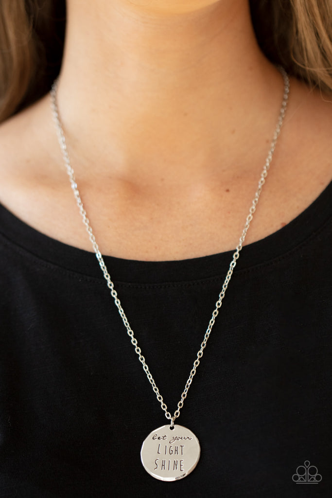 A shiny silver disc is stamped in the inspiring phrase, "Let your light shine," at the bottom of a shiny silver chain. Features an adjustable clasp closure.  Sold as one individual necklace. Includes one pair of matching earrings.