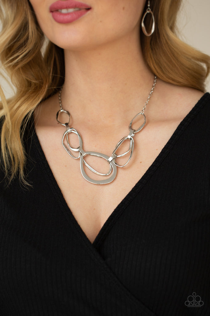 An asymmetrical assortment of twisted and flattened silver rings delicately link below the collar with oversized silver fittings, creating an intense industrial display below the collar. Features an adjustable clasp closure.  Sold as one individual necklace. Includes one pair of matching earrings.  