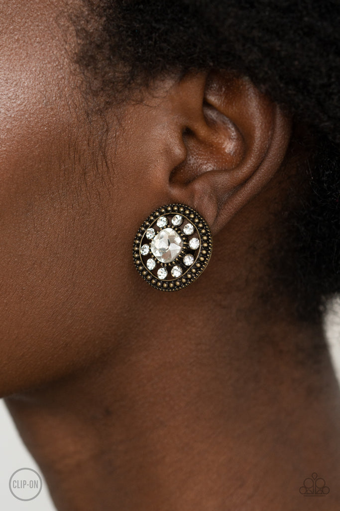 Featuring an oversized center, a white rhinestone encrusted flower blooms across the brass center of a studded frame for a sparkly finish. Earring attaches to a standard clip-on fitting.  Sold as one pair of clip-on earrings.