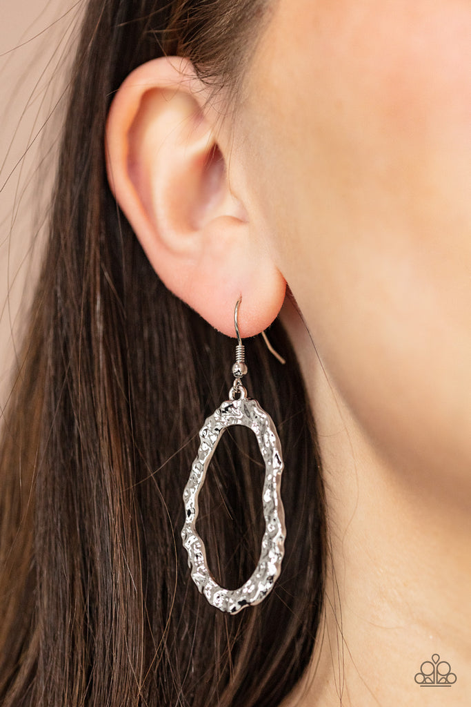 A high sheen silver oval frame is hammered into an asymmetrical frame, creating an edgy display. Earring attaches to a standard fishhook fitting.  Sold as one pair of earrings.  