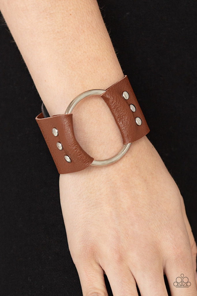 An oversized silver hoop is studded in place between the center of studded pieces of black and brown leather, creating a rustic centerpiece around the wrist. Features an adjustable snap closure.  Sold as one individual bracelet.  