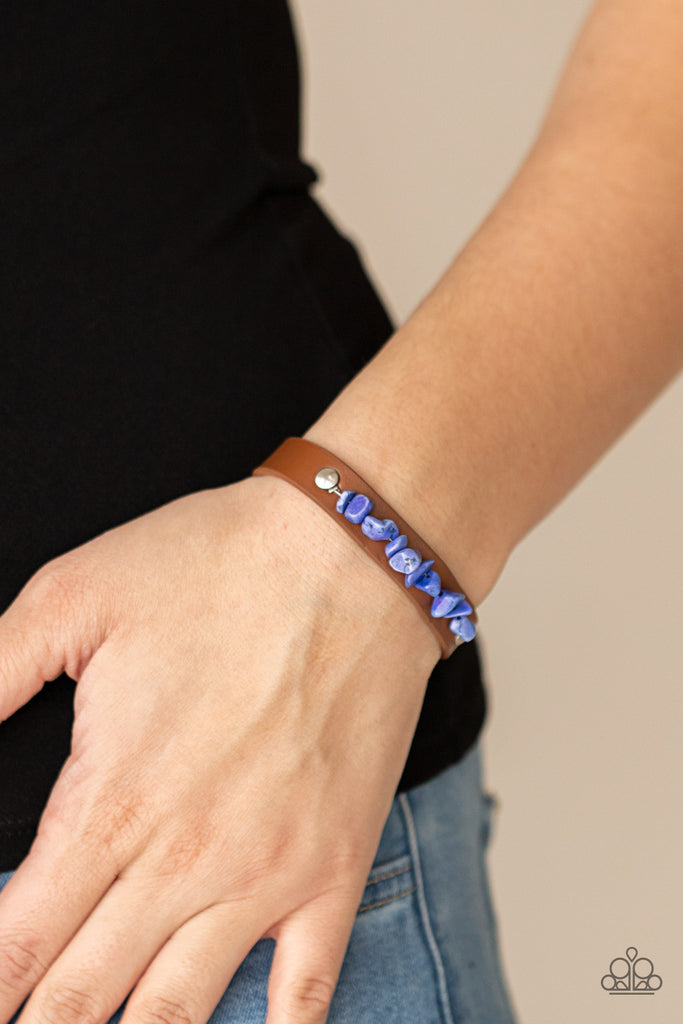 A strand of blue pebbles is studded in place across the front of a skinny brown leather band, creating a colorful earthy display around the wrist. Features an adjustable snap closure.  Sold as one individual bracelet.