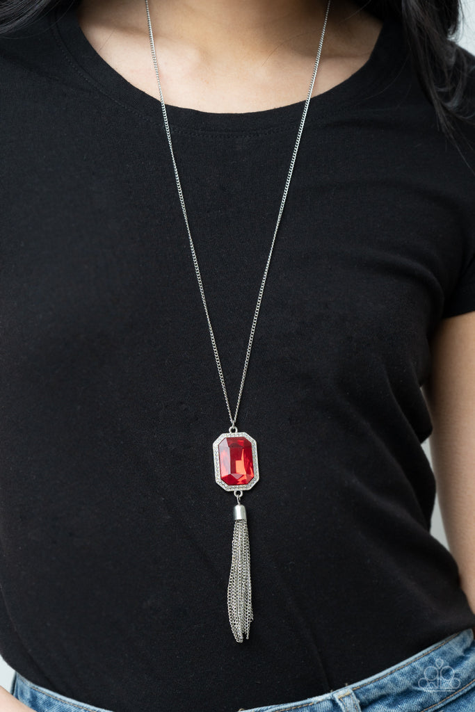 An impressive red emerald cut gem is pressed into the center of a silver studded frame, creating an ethereal pop of color at the bottom of a lengthened silver chain. A silver chain tassel swings from the bottom of the pendant, adding flirtatious movement to the opulent display. Features an adjustable clasp closure.  Sold as one individual necklace. Includes one pair of matching earrings.