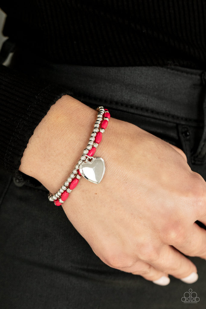 A bright silver heart dangles from a strand of playful Raspberry Sorbet beads. It is paired with a strand of round silver beads threaded along a stretchy band for a whimsical display around the wrist.  Sold as one individual bracelet.