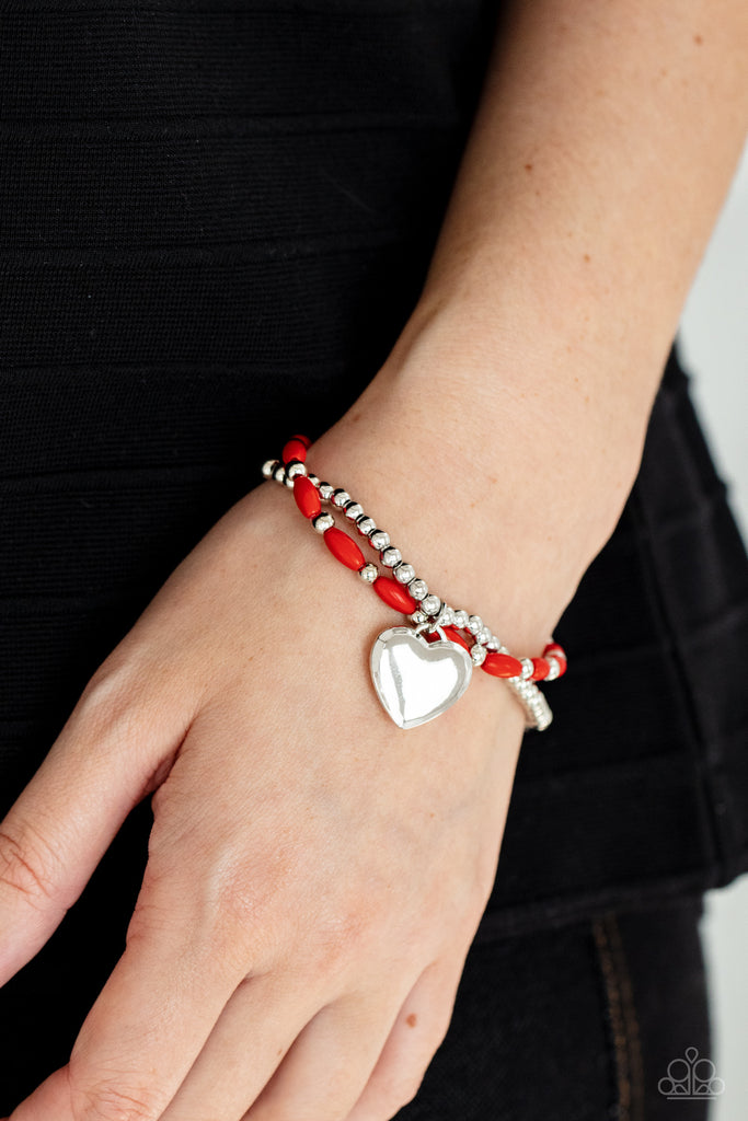 A shiny silver heart dangles from a strand of fiery red beads. It is paired with a strand of round silver beads threaded along a stretchy band for a whimsical display around the wrist.  Sold as one pair of bracelets.