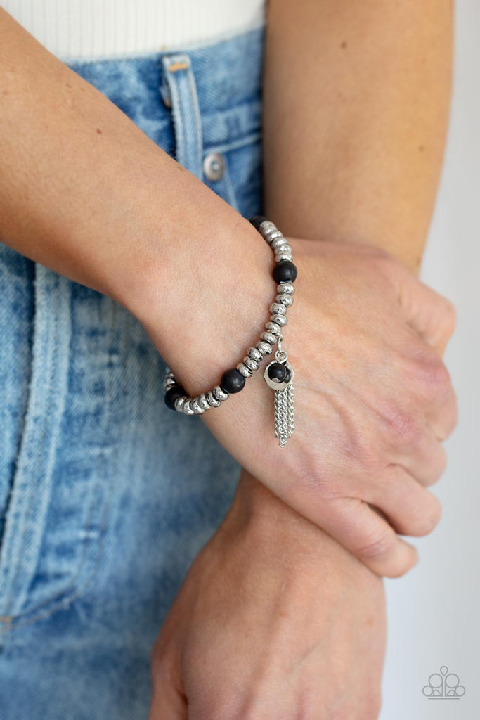 An earthy collection of hammered silver beads and round black stones are threaded along a stretchy band around the wrist. A dainty silver tassel, hammered silver disc, and a black stone swing from the seasonal display, adding playful movement to the whimsy look.  Sold as one individual bracelet.  