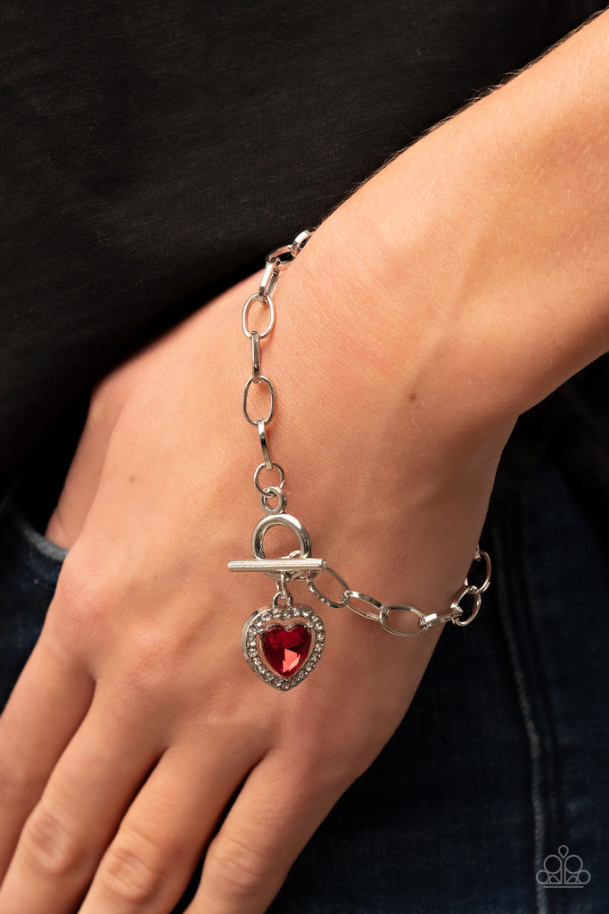Bordered in glassy white rhinestones, a red heart shaped gem sparkles from a toggle closure at the center of an oval silver linked chain for a flirtatious fashion around the wrist. Features a toggle closure.  Sold as one individual bracelet.  