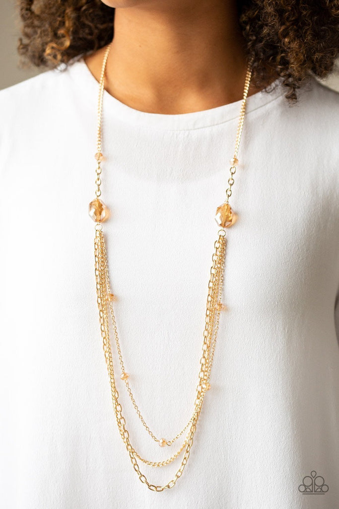 Infused with golden crystal-like beads, a pair of glittery golden gems gives way to layers of mismatched gold  chains for a dazzling look. Features an adjustable clasp closure.  Sold as one individual necklace. Includes one pair of matching earrings.