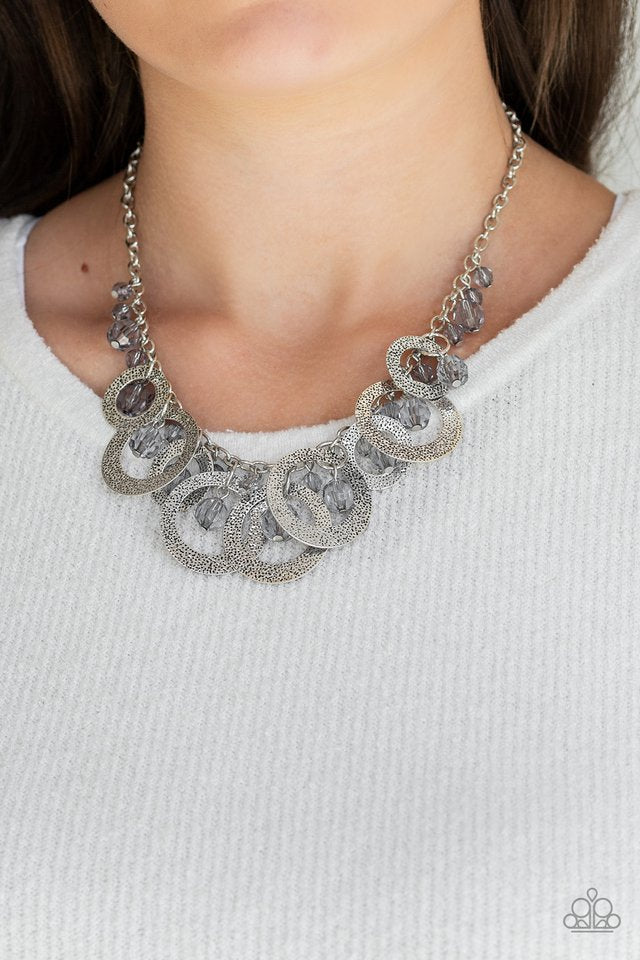 A collection of smoky crystal-like beads and hammered silver hoops dangle from the bottom of a shimmery silver chain, creating a noise-making fringe. Features an adjustable clasp closure.  Sold as one individual necklace. Includes one pair of matching earrings.