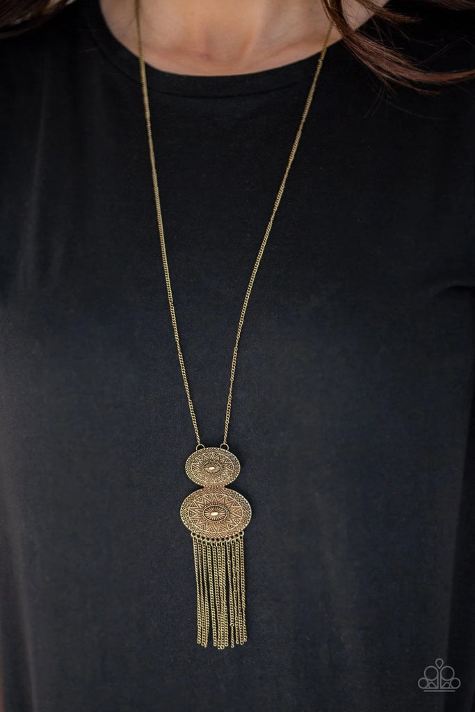 Embossed in radiant sunburst patterns, two oval brass frames connect at the bottom of a lengthened brass chain. Glistening brass chains stream from the bottom of the stacked pendant, adding a wanderlust flair to the tribal inspired look. Features an adjustable clasp closure.  Sold as one individual necklace. Includes one pair of matching earrings.