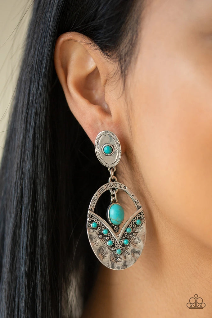 Rippling with hammered silver textures, a turquoise stone dotted frame swings from the bottom of an ornate oval frame dotted with a matching turquoise stone. A smooth turquoise stone swings from the top of the ornate frame, creating an earthy lure. Earring attaches to a standard post fitting.  Sold as one pair of post earrings.