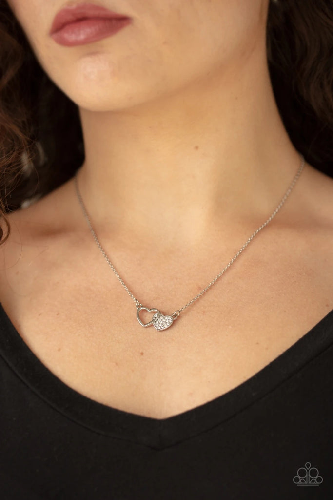 A white rhinestone encrusted silver heart joins a glistening silver heart silhouette at the bottom of a shimmery silver chain, creating a charming pendant below the collar. Features an adjustable clasp closure.  Sold as one individual necklace. Includes one pair of matching earrings.