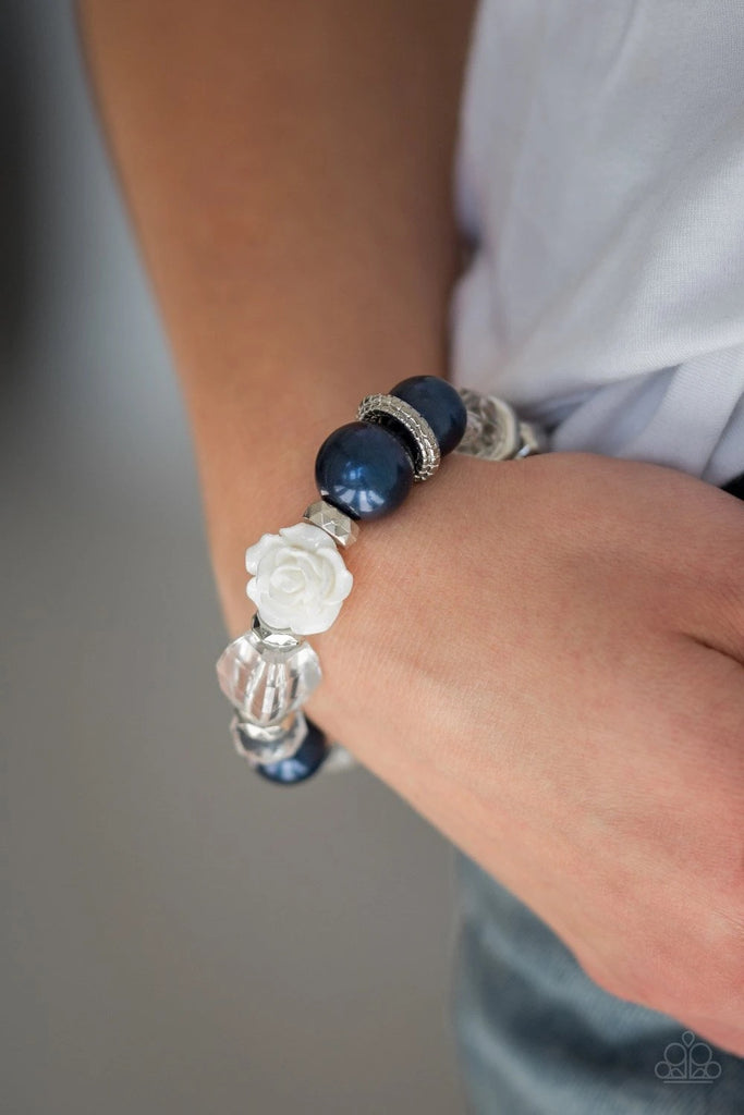 A collection of silver, pearly blue, and white crystal-like beads are threaded along a stretchy band around the wrist. A shiny white resin rose joins the refined palette for a glamorous finish.  Sold as one individual bracelet.